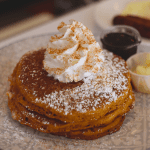Pumpkin pancakes topped with whipped cream and cinnamon sugar served with butter and maple syrup with eggs and sausage in the background
