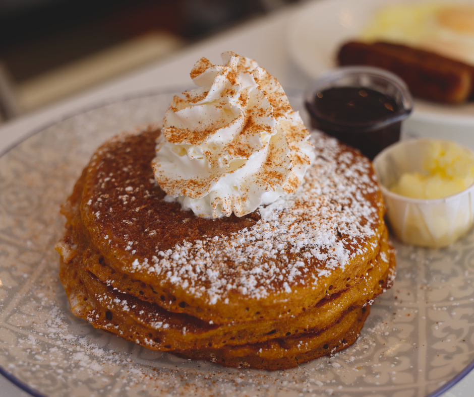Pumpkin pancakes topped with whipped cream and cinnamon sugar served with butter and maple syrup with eggs and sausage in the background